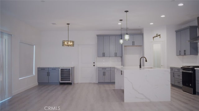 kitchen featuring light hardwood / wood-style floors, beverage cooler, decorative light fixtures, stainless steel stove, and an island with sink