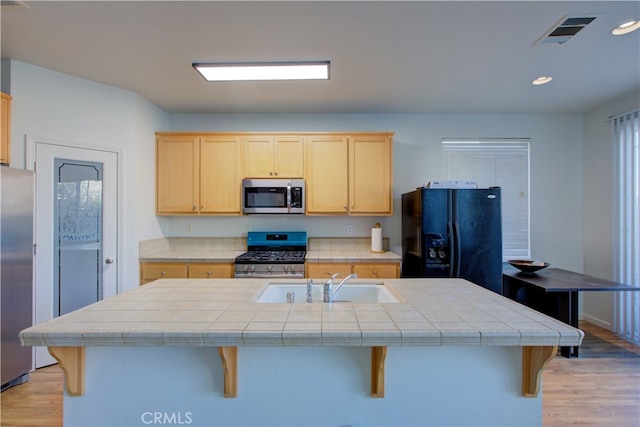 kitchen with light brown cabinetry, sink, tile countertops, stainless steel appliances, and a center island with sink