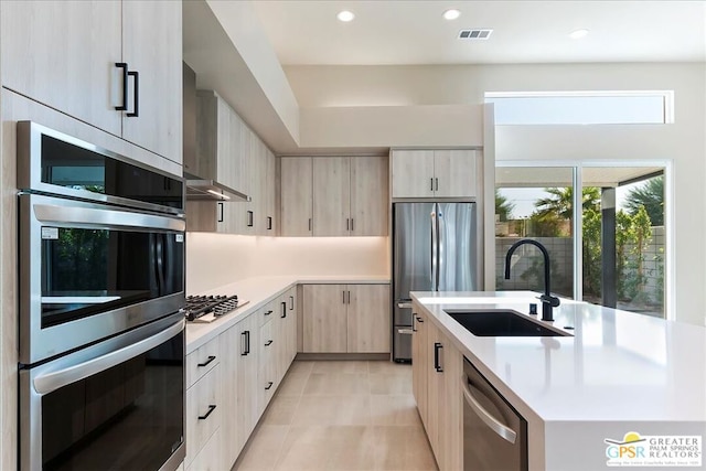 kitchen with light brown cabinetry, wall chimney range hood, sink, an island with sink, and stainless steel appliances