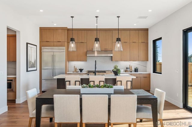 kitchen with light hardwood / wood-style flooring, decorative light fixtures, stainless steel appliances, backsplash, and a center island with sink