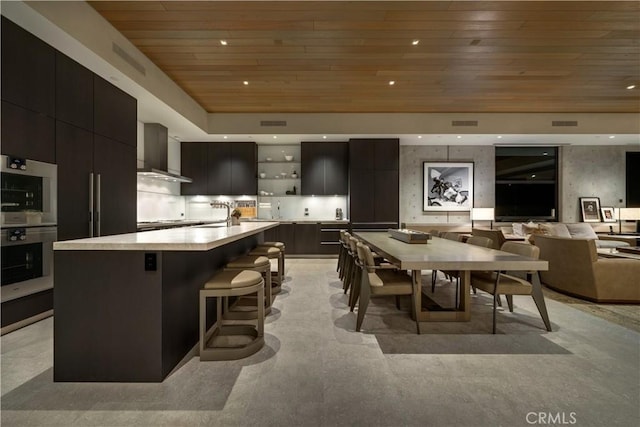 kitchen featuring a breakfast bar, wooden ceiling, a kitchen island with sink, multiple ovens, and wall chimney exhaust hood