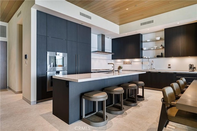 kitchen with a kitchen breakfast bar, wall chimney range hood, double wall oven, an island with sink, and wood ceiling