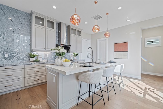 kitchen with light hardwood / wood-style flooring, wall chimney exhaust hood, a kitchen island with sink, and pendant lighting