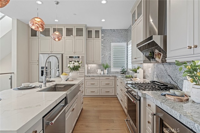 kitchen with wall chimney range hood, hanging light fixtures, light hardwood / wood-style floors, stainless steel appliances, and decorative backsplash