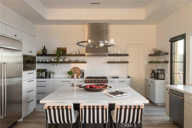 kitchen featuring built in appliances, island range hood, white cabinets, and a kitchen island with sink