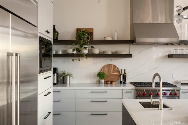 kitchen featuring built in appliances, wall chimney exhaust hood, white cabinetry, and backsplash