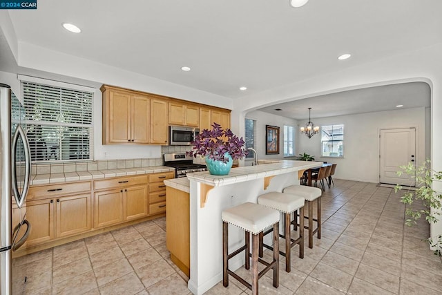 kitchen featuring light tile patterned flooring, hanging light fixtures, stainless steel appliances, tile counters, and a center island with sink