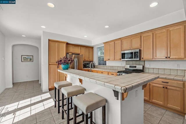 kitchen featuring a kitchen bar, tile countertops, stainless steel appliances, light tile patterned floors, and a center island with sink