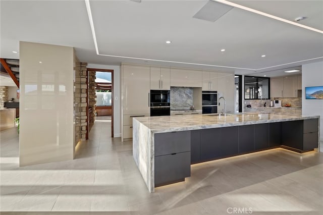 kitchen with decorative backsplash, a large island with sink, light tile patterned flooring, gray cabinets, and double oven