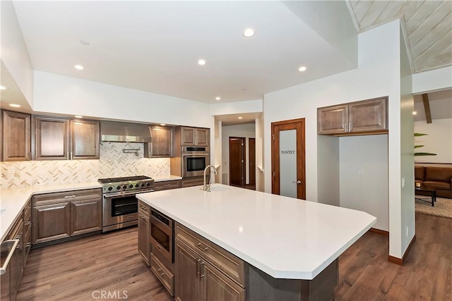 kitchen with appliances with stainless steel finishes, tasteful backsplash, wall chimney exhaust hood, a kitchen island with sink, and dark wood-type flooring