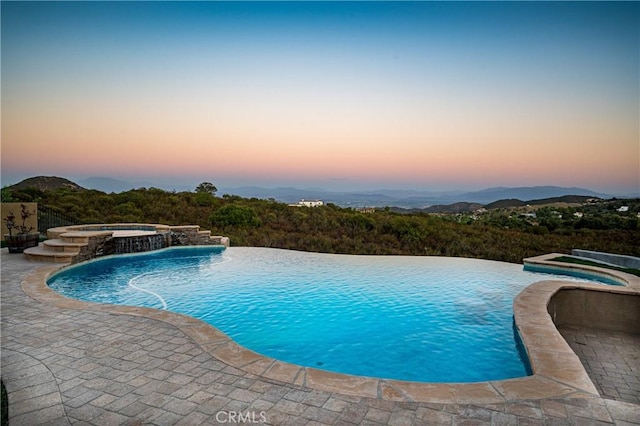 pool at dusk featuring pool water feature, a mountain view, a patio area, and an in ground hot tub