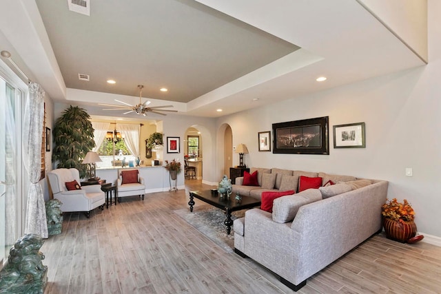 living room with a raised ceiling, ceiling fan, and light wood-type flooring