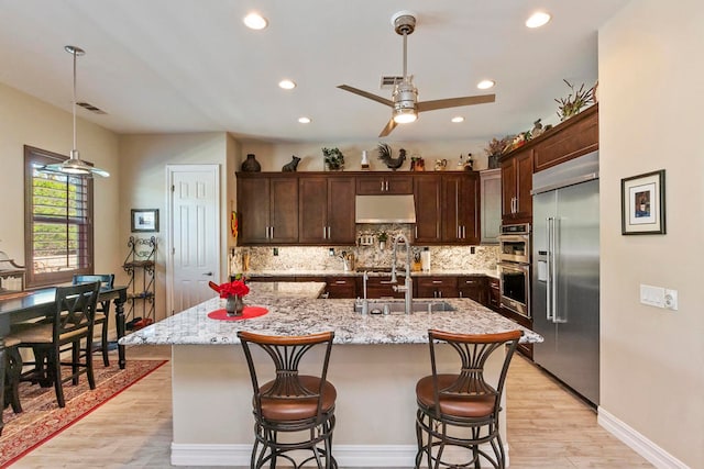 kitchen with light stone countertops, appliances with stainless steel finishes, light wood-type flooring, sink, and a center island with sink