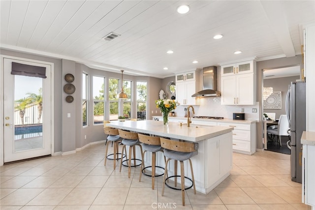 kitchen featuring white cabinets, wall chimney range hood, tasteful backsplash, an island with sink, and stainless steel refrigerator