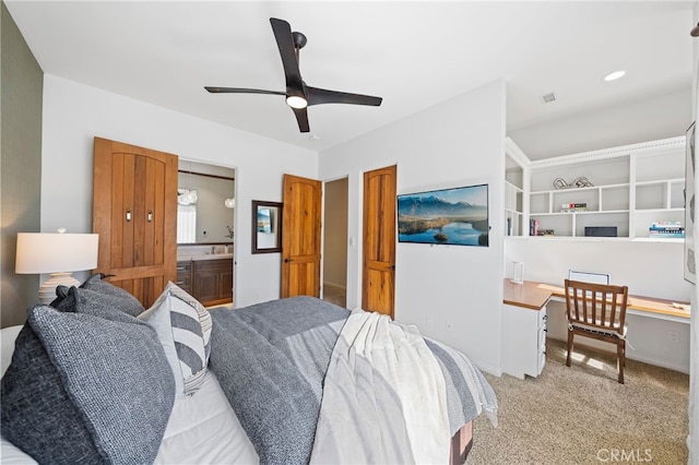 bedroom featuring built in desk, ensuite bath, light colored carpet, and ceiling fan