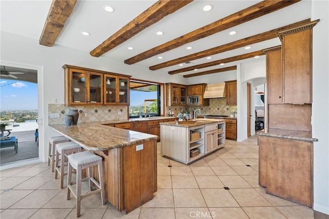 kitchen with stainless steel microwave, sink, backsplash, beam ceiling, and a kitchen island with sink