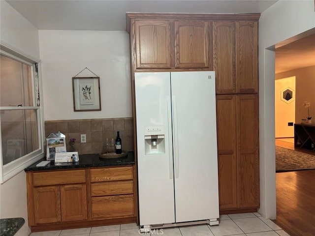 kitchen with light tile patterned floors, backsplash, white refrigerator with ice dispenser, and dark stone countertops