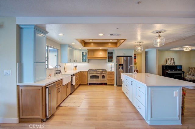 kitchen with light hardwood / wood-style flooring, a tray ceiling, a center island with sink, pendant lighting, and appliances with stainless steel finishes