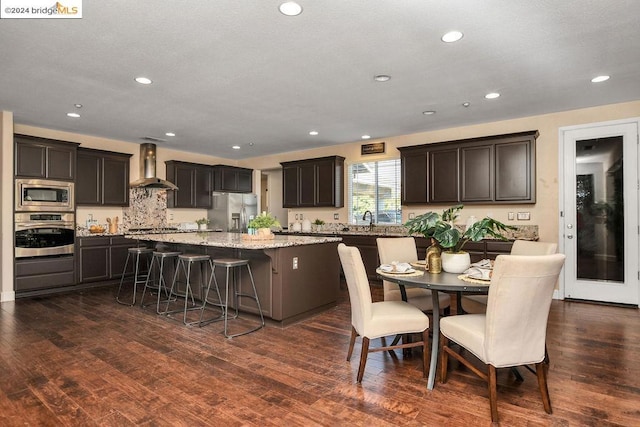 kitchen featuring wall chimney range hood, dark hardwood / wood-style flooring, stainless steel appliances, dark brown cabinetry, and a center island