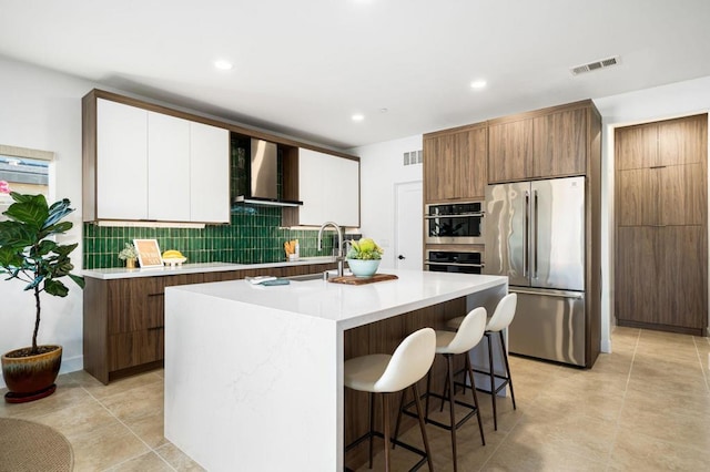 kitchen with white cabinetry, wall chimney exhaust hood, decorative backsplash, a center island with sink, and appliances with stainless steel finishes