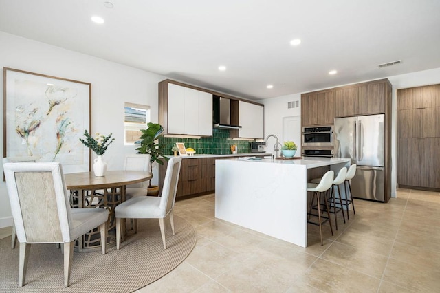 kitchen featuring backsplash, wall chimney exhaust hood, stainless steel appliances, a kitchen island with sink, and light tile patterned flooring