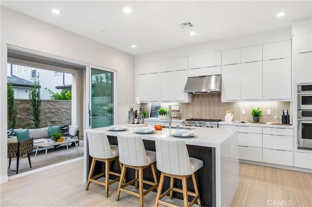 kitchen with white cabinets, an island with sink, light wood-type flooring, and ventilation hood