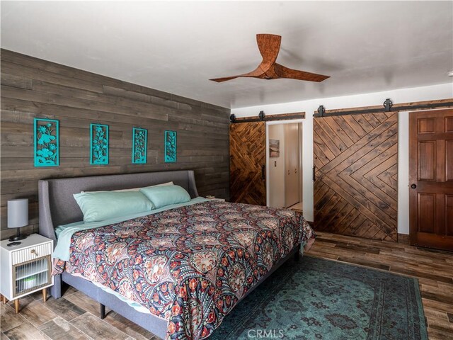 bedroom with a barn door, ceiling fan, wooden walls, and hardwood / wood-style flooring