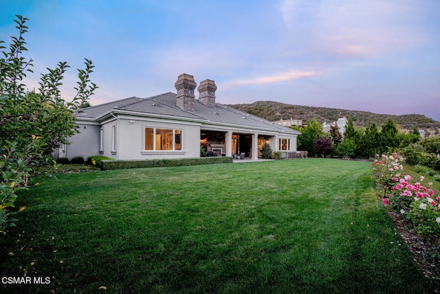 back house at dusk featuring a mountain view, a patio area, and a yard