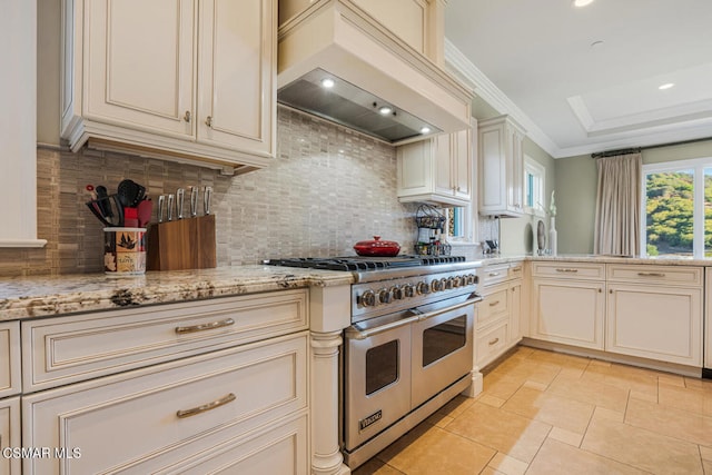 kitchen featuring range with two ovens, backsplash, cream cabinets, light stone counters, and custom range hood