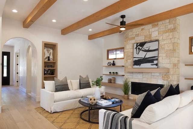 living room with beamed ceiling, light hardwood / wood-style flooring, a stone fireplace, and ceiling fan