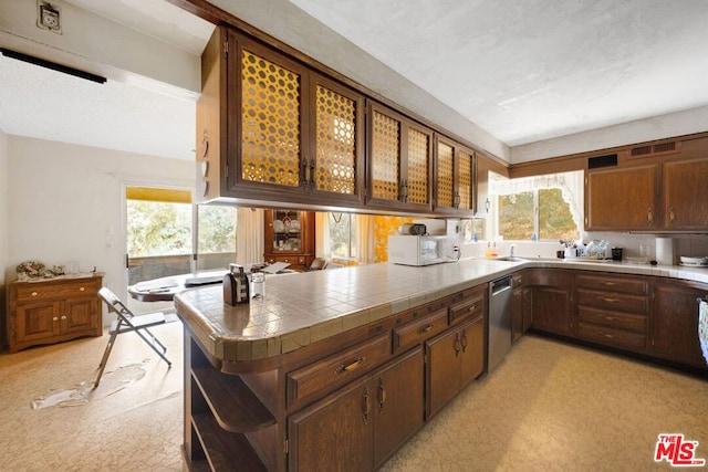 kitchen with stainless steel dishwasher, dark brown cabinetry, kitchen peninsula, and tile counters