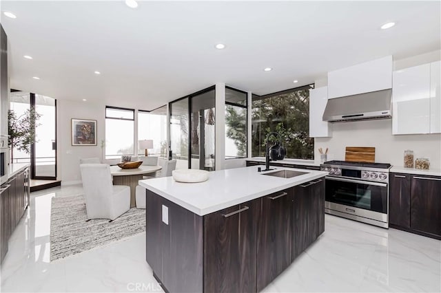 kitchen with stainless steel stove, sink, white cabinets, a kitchen island with sink, and wall chimney range hood