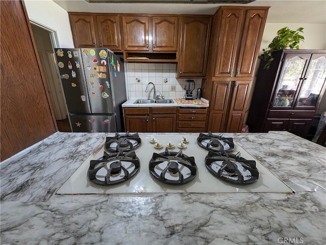 kitchen with white gas cooktop, sink, decorative backsplash, stainless steel fridge, and light stone counters