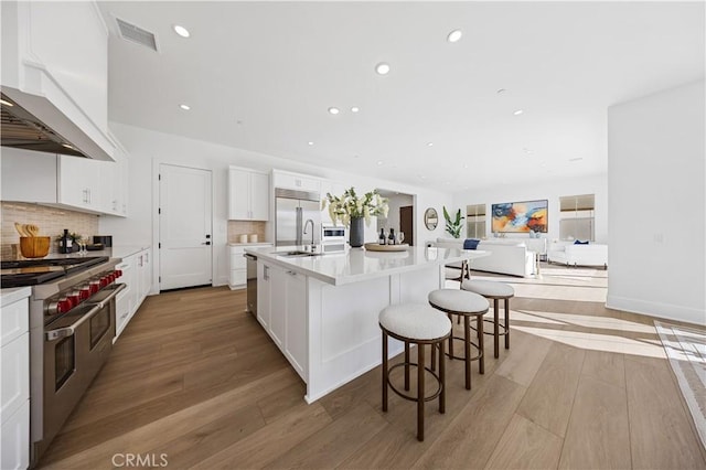 kitchen featuring white cabinets, wall chimney exhaust hood, high end appliances, and an island with sink