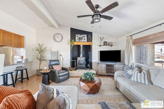 living room featuring light wood-type flooring, lofted ceiling with beams, a wood stove, a textured ceiling, and ceiling fan