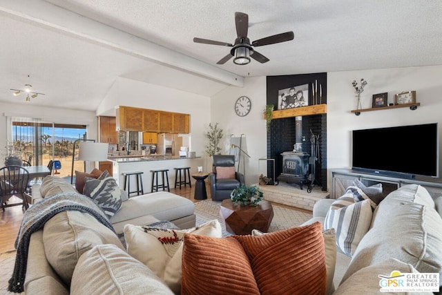 living room with vaulted ceiling with beams, a textured ceiling, a wood stove, ceiling fan, and light hardwood / wood-style flooring