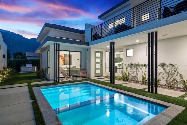 back house at dusk featuring a patio area, a balcony, a mountain view, a lawn, and a swimming pool with hot tub