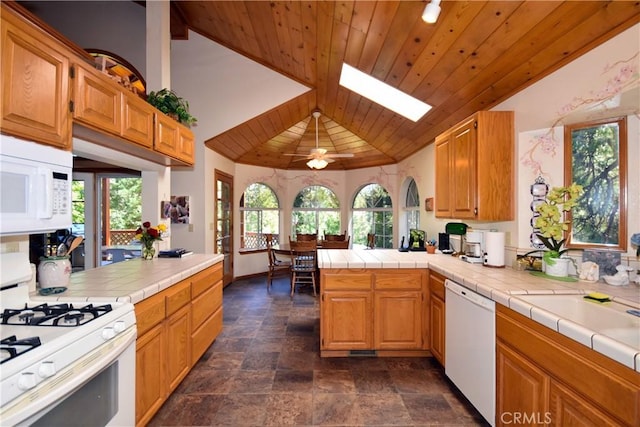 kitchen featuring white appliances, stone finish floor, a peninsula, tile countertops, and lofted ceiling with skylight