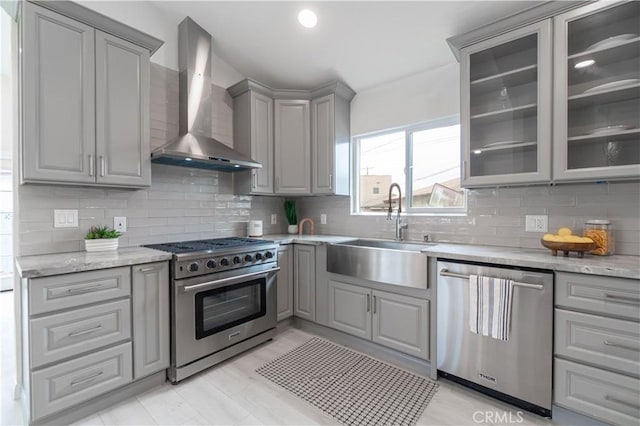 kitchen with stainless steel appliances, a sink, wall chimney range hood, gray cabinets, and glass insert cabinets