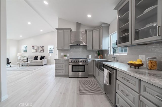 kitchen featuring light wood-style flooring, gray cabinetry, appliances with stainless steel finishes, vaulted ceiling, and wall chimney exhaust hood