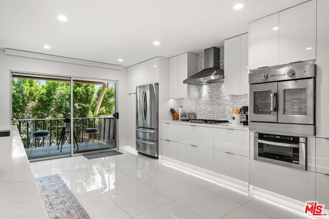 kitchen with light stone countertops, wall chimney range hood, white cabinetry, stainless steel appliances, and decorative backsplash