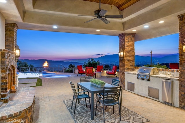 patio terrace at dusk featuring area for grilling, a swimming pool, exterior kitchen, ceiling fan, and a mountain view