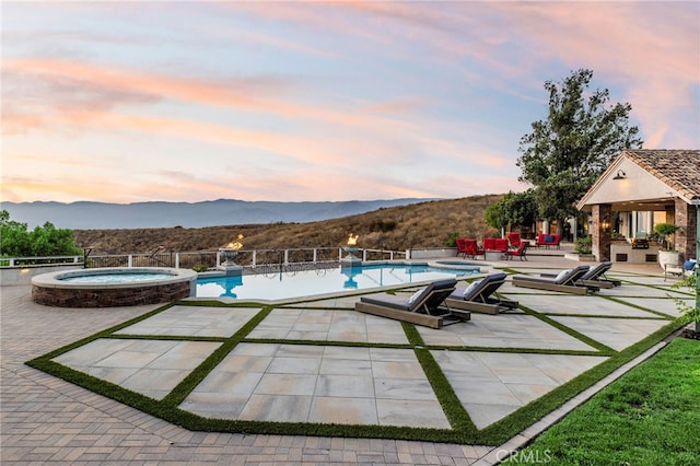 pool at dusk with an in ground hot tub, a mountain view, and a patio