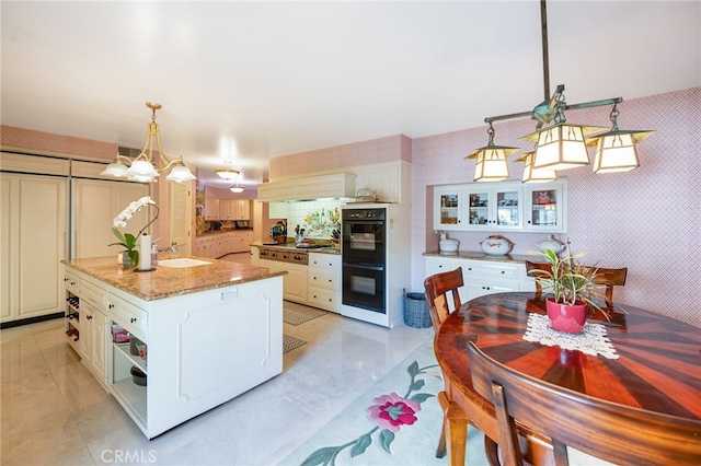 kitchen with black double oven, light stone counters, hanging light fixtures, and a kitchen island with sink