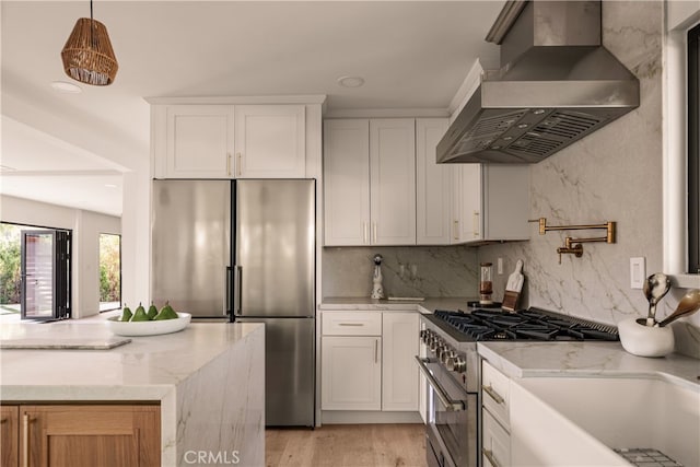 kitchen with wall chimney exhaust hood, light stone countertops, white cabinetry, appliances with stainless steel finishes, and light hardwood / wood-style floors