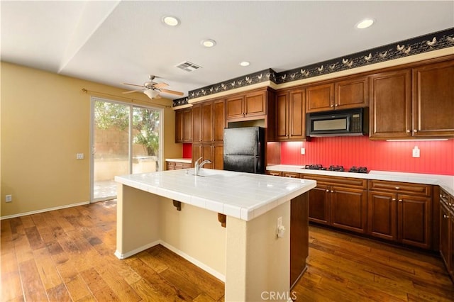 kitchen with ceiling fan, dark hardwood / wood-style floors, tile countertops, an island with sink, and black appliances