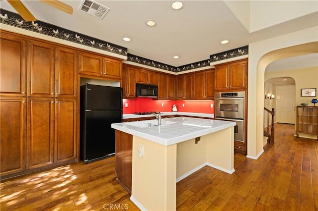 kitchen featuring ceiling fan, black appliances, tile countertops, dark hardwood / wood-style floors, and an island with sink