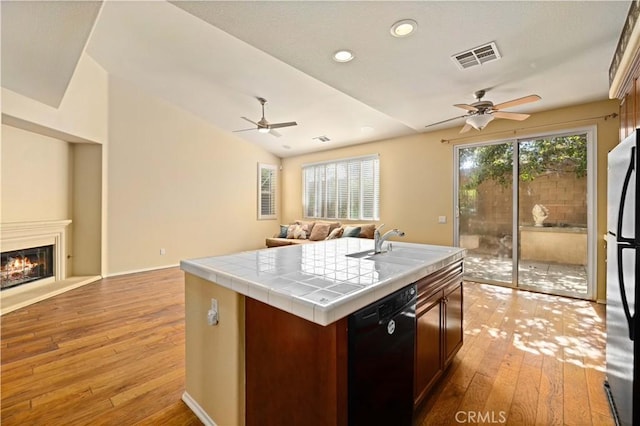 kitchen featuring dishwasher, tile countertops, stainless steel fridge, light hardwood / wood-style floors, and a center island with sink