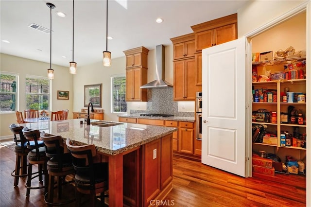 kitchen with light stone countertops, wall chimney range hood, sink, hanging light fixtures, and a center island with sink