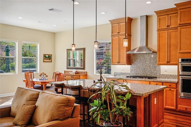 kitchen featuring wall chimney range hood, a kitchen island, pendant lighting, light stone countertops, and stainless steel double oven